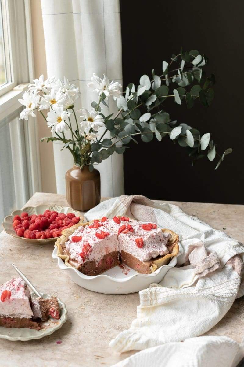 Raspberry mocha french silk pie stylized with a linen napkin, a plate that has a slice of the pie that has been cut in to, a plate of fresh raspberries, and flowers.